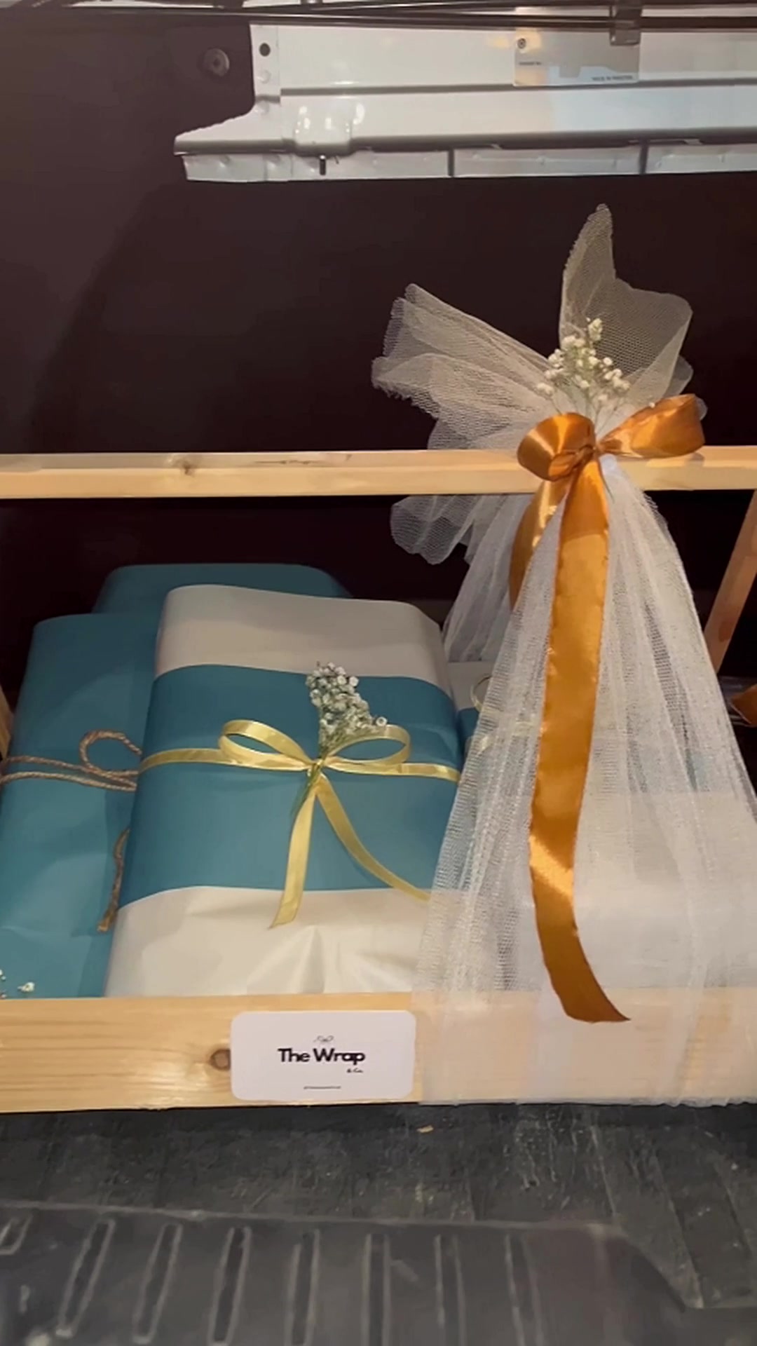 Wrapped gifts arranged in a wooden crate with teal and ivory paper, satin ribbons and The Wrap & Co. card